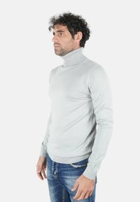 Maglione dolcevita grigio chiaro in tessuto morbido, con collo a costine e maniche lunghe. Indossato con jeans blu.