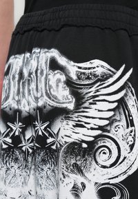 Μαύρο παντελόνι φόρμας με έντονο λευκό γραφικό σχέδιο, που περιλαμβάνει φτερά, σπειροειδή μοτίβα και αστέρια στην περιοχή του μηρού.