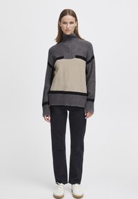 Un pull gris et beige avec un col montant, comportant des rayures horizontales noires. Porté avec un pantalon noir et des baskets blanches. Texture lisse.