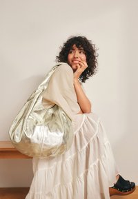 Metalna zlatna hobo torba iz kože, sa zaobljenim oblikom, glatkom teksturom i širokom naramenicom. Nošena ležerno na svijetloj pozadini.