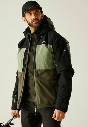 Mężczyzna w czarnej czapce i zielono-czarnej kurtce, trzymający kijki trekkingowe, patrzący lekko w górę i na bok na jednolitym tle.