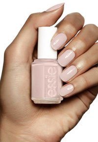 Flacon de vernis à ongles Essie dans une teinte rose pâle tenu dans une main avec des ongles assortis très bien manucurés. Le flacon présente un bouchon blanc et une étiquette texturée.