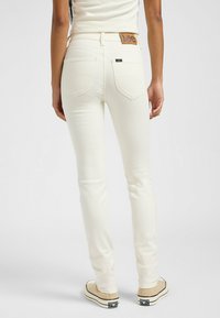 Jeans blancos con un ajuste slim, bolsillos estándar y una etiqueta de cuero con la marca en la parte trasera. Combinados con calzado de color marrón claro.