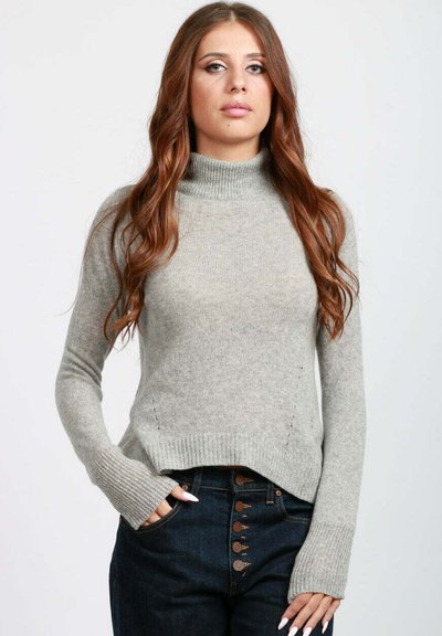 Donna con lunghi capelli mossi castani che indossa un maglione grigio chiaro a collo alto e jeans a vita alta blu scuro con bottoni decorativi.