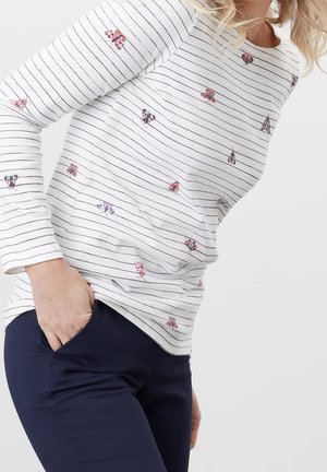 Femme portant un haut blanc à manches longues avec de fines rayures noires et de petites impressions florales colorées, associé à un pantalon bleu foncé.