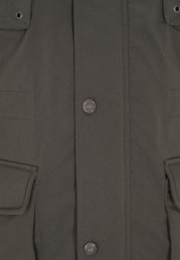 Giacca verde scuro in tessuto testurizzato, chiusura frontale con bottoni, due grandi tasche a patta e una toppa rotonda con logo sul lato sinistro.