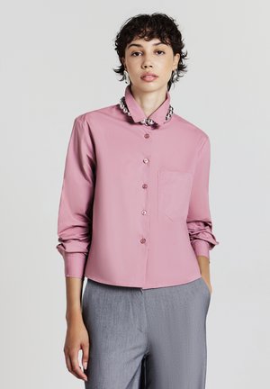 Jeune personne avec des cheveux bouclés courts portant une chemise rose à boutons avec des perles argentées au col et un pantalon gris, se tenant devant un fond uni.