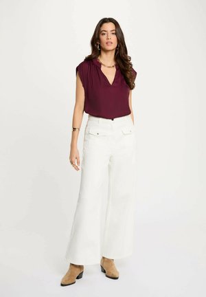 Donna che indossa una blusa bordeaux senza maniche, pantaloni bianchi a gamba larga a vita alta con tasche anteriori, stivaletti color cuoio, gioielli in oro, in piedi su sfondo bianco.