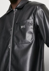 Zwarte korte jas van nep leer met korte mouwen, een ritssluiting aan de voorkant, een puntige kraag en een opgestikt zakje met een metalen logodetail. Gladde textuur.