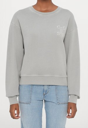 Personne portant un sweat-shirt gris clair à col rond avec le texte blanc « CLOSED » sur la poitrine et un jean bleu clair, debout devant un fond uni.