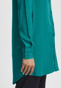 Teal long-sleeve shirt made of smooth fabric. Features a button-up front and two-button cuffs, with a slightly curved hemline.