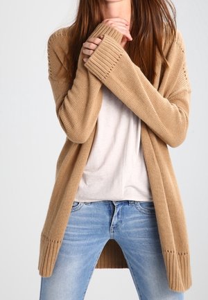 Femme aux longs cheveux bruns portant un cardigan beige en tricot par-dessus une chemise blanche et un jean bleu, les mains jointes près de la poitrine.
