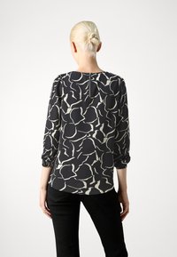 Femme aux cheveux blonds en chignon, portant un blouse à motifs abstraits noir et blanc et un pantalon noir, de dos contre un fond neutre.
