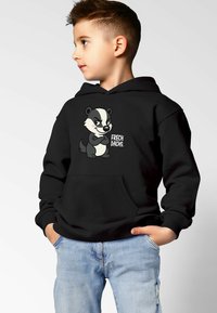 Junge trägt schwarzen Kapuzenpullover mit einem Cartoon-Dachs und dem Schriftzug "FRECH DACHS", kombiniert mit hellblauen Jeans, schaut seitlich und hat die Hände in den Taschen.