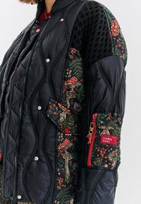 Pikowana czarna kurtka puchowa z akcentami w kwiatowy wzór, z panelami siateczkowymi, zatrzaskami oraz kieszenią na zamek błyskawiczny z czerwoną metką.