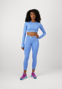 Svjetloplava Nike Pro majica s dugim rukavima i odgovarajuće tajice viskog struka, s akcentskim logotipima. Upareno s ljubičastim sportskim cipelama.