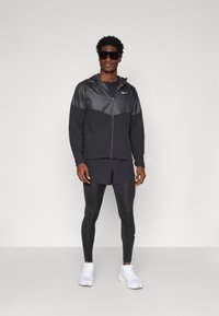 Czarna kurtka sportowa z błyszczącego materiału, dopasowane legginsy oraz białe sneakersy. Modelka nosi oversize'owe okulary przeciwsłoneczne. Równomierne oświetlenie, neutralne tło.