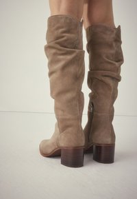 Bottes cuissardes en daim brun clair avec un design ample et une fermeture éclair latérale. Le talon épais présente une finition en bois texturé.