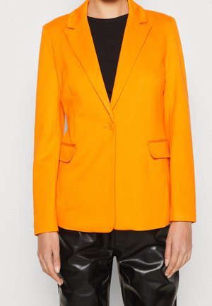 Kobieta ubrana w jasno pomarańczową marynarkę na jeden guzik na czarnym topie, zestawioną z błyszczącymi czarnymi skórzanymi spodniami, pokazana od ramion do bioder.