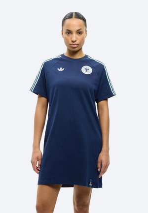 Kobieta w granatowej sukience t-shirt marki Adidas z białymi paskami na rękawach oraz logo sportowymi na piersi i u dołu, stojąca na jednolitym tle.