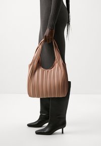 Borsa a spalla plissettata rosa pallido con texture morbida e manici larghi, tenuta da una persona in un vestito grigio a coste e stivali neri con tacco.