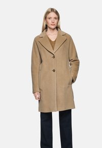 Frau in einem beigen, knielangen Wollmantel mit breiten Revers, zwei vorderen Knöpfen und Händen in den Taschen, kombiniert mit dunklen Hosen.