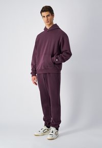 Paarse hoodie met een voorzak, gecombineerd met bijpassende joggingsbroek. Witte sneakers met zwarte en zilveren accenten maken de look af.