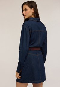 Jeansjurk met lange mouwen, donkerblauwe kleur, getailleerde pasvorm en een bruine riem. Bevat gouden knoopaccenten en zichtbare stiksels.