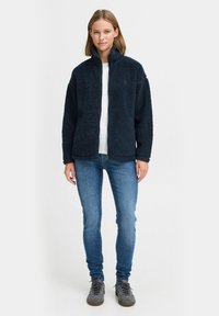 Veste en polaire bleu marine avec col haut, surface texturée et fermeture éclair à l'avant, associée à un jean ajusté bleu clair et des baskets grises.
