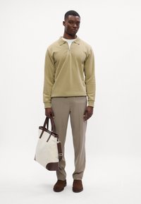 Beige cipzáras nyakú pulóver, világos barna nadrág, barna velúr cipők és egy krém színű táska barna bőrdíszítéssel. Egyszerű háttér.