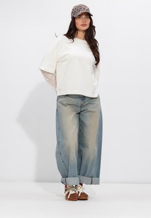 Woman standing wearing leopard print cap, loose white top over lace sleeves, wide cuffed jeans, and brown sneakers with white laces.