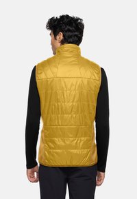 Gelbe Steppweste mit hohem Kragen, ausgestattet mit gesteppten Details und seitlichen Einsätzen. Über einem schwarzen Langarmshirt getragen.