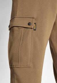 Bruna cargobyxor med en sidficka med klaff, två svarta knappar och en liten gul och svart 'BOSS'-etikett. Slät textur.