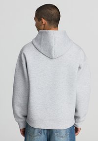 Felpa grigia con cappuccio, in tessuto texturizzato, taglio oversize e polsini a coste, abbinata a jeans blu chiaro. Vista da dietro.