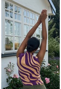 Top sans manches en tricot présentant des rayures ondulées alternées violet et pêche, col montant et tissu texturé. Arrière-plan avec des roses.