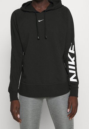 Personne portant un sweat à capuche noir Nike avec un logo blanc sur la poitrine et la manche, assorti à des leggings gris clair.