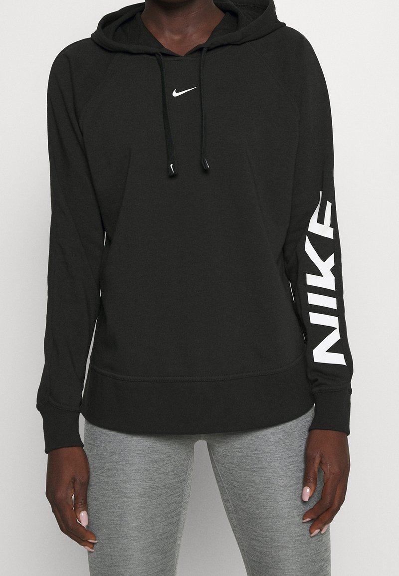 Personne portant un sweat à capuche noir Nike avec un logo blanc sur la poitrine et la manche, assorti à des leggings gris clair.