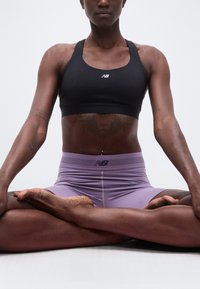 Άτομο καθισμένο με σταυρωμένα πόδια στο πάτωμα, φορώντας μαύρο αθλητικό σουτιέν και μοβ σορτς, ορατός κορμός και χέρια με τατουάζ, απλό ανοιχτόχρωμο φόντο.