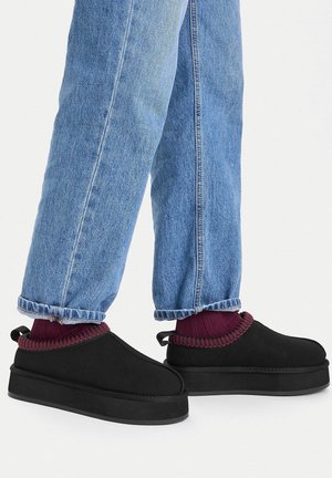 Zapatos negros sin cordones con suela gruesa y exterior suave, con un ribete acanalado color granate en el tobillo, combinados con vaqueros azules con puños.