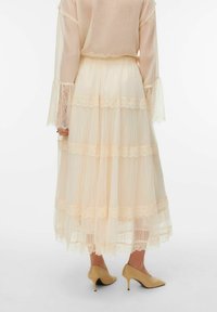 Jupe longue à plusieurs couches en tissu pêche doux avec bordure en dentelle. Texture transparente et forme évasée, associée à des chaussures à talons hauts beiges.