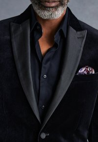 Blazer de terciopelo negro con solapas de satén, combinado con una camisa negra texturizada. Presenta un pañuelo de bolsillo con un patrón colorido en el bolsillo del pecho.