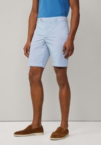 Shorts bleu clair avec un devant plat, des passants de ceinture et des poches latérales. Fabriqués en coton avec une texture lisse. Chaussures slip-on en daim marron.