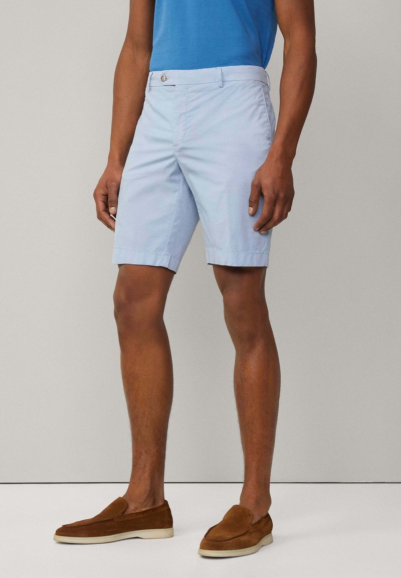 Shorts bleu clair avec un devant plat, des passants de ceinture et des poches latérales. Fabriqués en coton avec une texture lisse. Chaussures slip-on en daim marron.