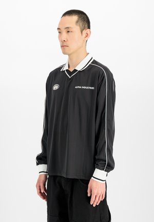 Jonge man draagt een zwart sportshirt met lange mouwen met witte strepen en Alpha Industries-logo op een witte achtergrond.