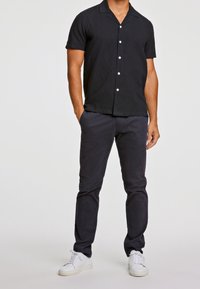 Camicia nera a maniche corte con bottoni bianchi, abbinata a pantaloni scuri e sneakers bianche, che presenta un look casual e aderente.
