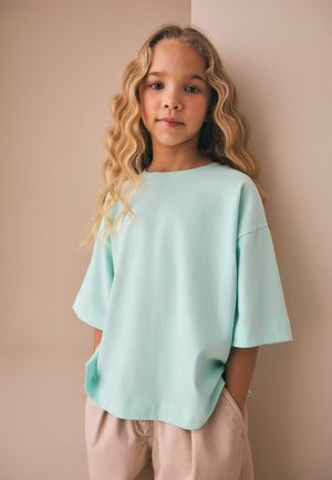 Junges Mädchen mit langen, welligen blonden Haaren, das ein hellblaues übergroßes T-Shirt und beige Hose trägt, steht vor einer beigen Wand.