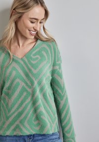 Uśmiechnięta kobieta o blond włosach nosi zielony i beżowy wzorzysty, miękki sweter oraz niebieskie dżinsy, patrząc w dół na jednolitym szarym tle.