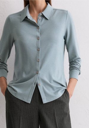 Kobieta ubrana w jasnoniebieską, zapinaną na guziki koszulę z długim rękawem i kołnierzykiem, lekko włożoną do ciemnych spodni w prążki, z rękami w kieszeniach.