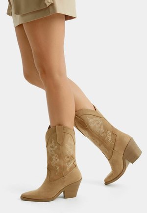 Jambes portant des bottes de cowboy en daim beige avec des motifs décoratifs brodés et des talons carrés, associées à une jupe beige sur un fond blanc.