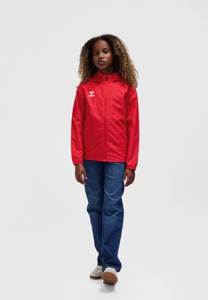 Rød vandtæt jakke med hætte, lynlås og sidelommer. Kombineret med blå jeans og lyse sneakers. Har logo på brystet.